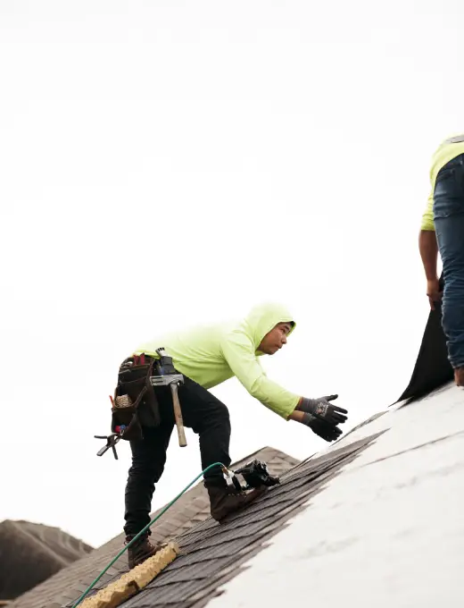 A roofer in a yellow hoodie installing shingles on a residential roof.