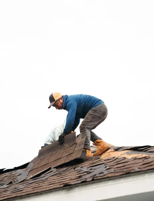 A worker wearing a hat and gloves removes shingles from a roof